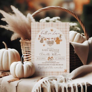 May include: A baby shower invitation with a fall theme. The invitation features a plaid background and an arch with hanging baby clothes and a teddy bear. The text reads "Fall in Love" and "Baby Shower". Pumpkins and a basket are in the background.