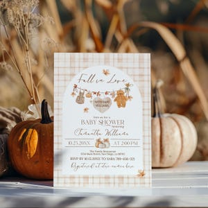 May include: Baby shower invitation with a fall theme. The invitation features the text "Fall in Love" and "Baby Shower" with decorative elements like leaves and a teddy bear. The background has a plaid pattern.