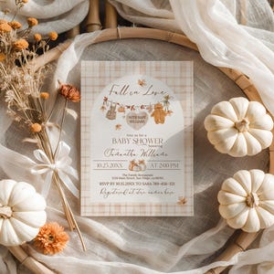 May include: A baby shower invitation with a fall theme. The invitation features the text "Fall in Love" and "Baby Shower" with decorative elements like pumpkins and leaves. The invitation is surrounded by white pumpkins, dried flowers, and fabric.