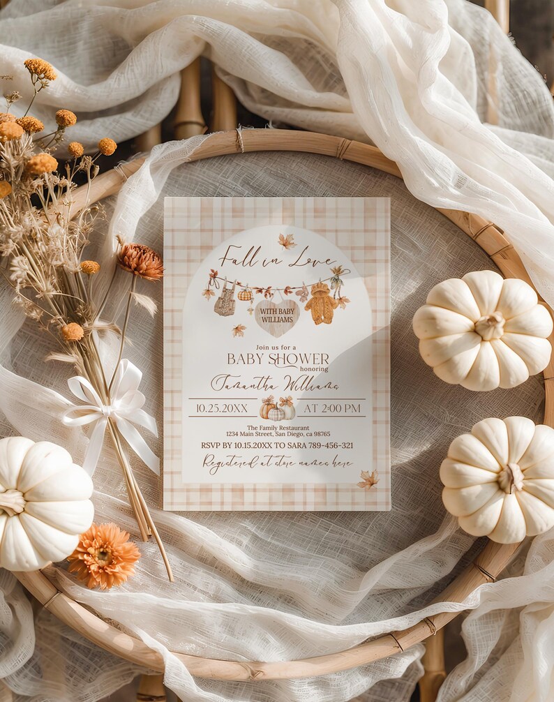 May include: A baby shower invitation with a fall theme. The invitation features the text "Fall in Love" and "Baby Shower" with decorative elements like pumpkins and leaves. The invitation is surrounded by white pumpkins, dried flowers, and fabric.