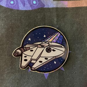 May include: A holographic sticker of a white spaceship flying through a dark blue space with stars and a sparkly trail.
