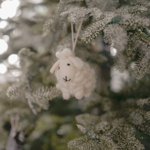 May include: A white felt sheep ornament hanging on a snowy artificial Christmas tree.
