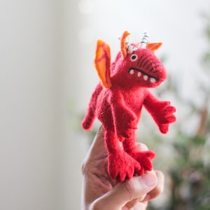 May include: A handmade red felt dragon toy with orange wings and white horns. The dragon has a detailed face with white teeth and black stitching. The toy is held up by a hand.