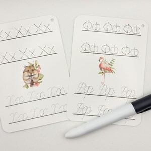 May include: Two white dry-erase boards with dotted lines for practicing handwriting. One board features a hamster illustration and the other features a flamingo illustration. The boards have holes in the top corner for hanging. A black and white dry-erase marker is included.