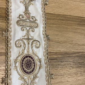 May include: A white fabric trim featuring an intricate embroidered pattern in metallic gold thread and deep purple accents. The design includes repeating oval medallions and scrolling floral motifs, finished with a scalloped gold edge trim.
