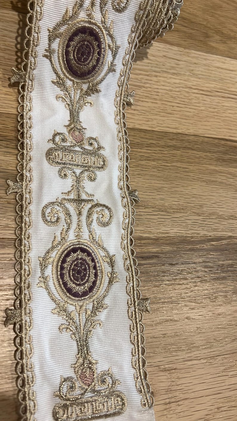 May include: A white fabric trim featuring an intricate embroidered pattern in metallic gold thread and deep purple accents. The design includes repeating oval medallions and scrolling floral motifs, finished with a scalloped gold edge trim.