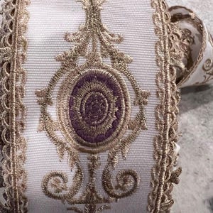 May include: A white fabric ribbon featuring intricate gold metallic embroidery and a central purple oval motif. The edges are finished with a decorative gold scalloped trim, and the design includes symmetrical scrollwork patterns throughout.