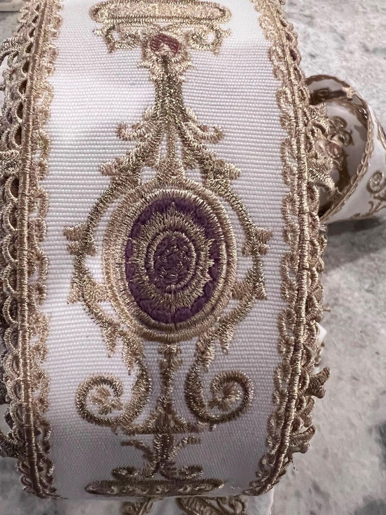 May include: A white fabric ribbon featuring intricate gold metallic embroidery and a central purple oval motif. The edges are finished with a decorative gold scalloped trim, and the design includes symmetrical scrollwork patterns throughout.