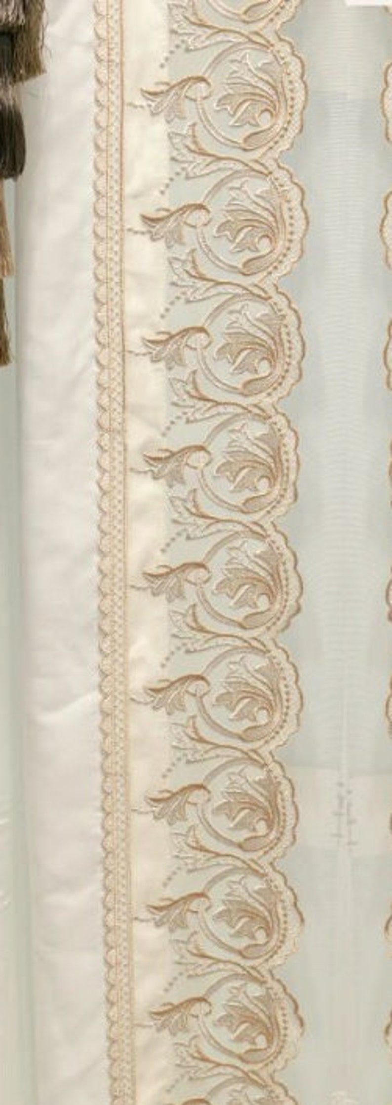 May include: Cream-colored curtain panel with intricate embroidered floral and scrollwork design in a light brown hue. The panel features a scalloped edge and a decorative tassel.