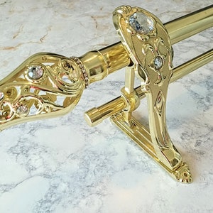 May include: Gold-toned curtain rod set with ornate, decorative finials and brackets. The finials feature clear crystal accents and intricate openwork designs. The rod is made of metal and is designed to hold curtains. The set includes two rods and two brackets.