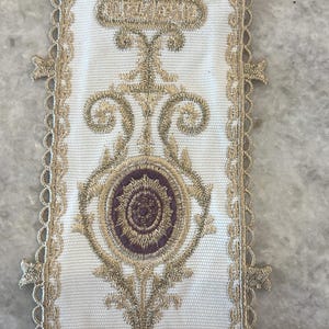 May include: A white fabric trim featuring intricate gold metallic embroidery and a central purple circular motif. The trim has scalloped edges and is positioned next to a metal ruler showing a width of approximately 3.5 inches.