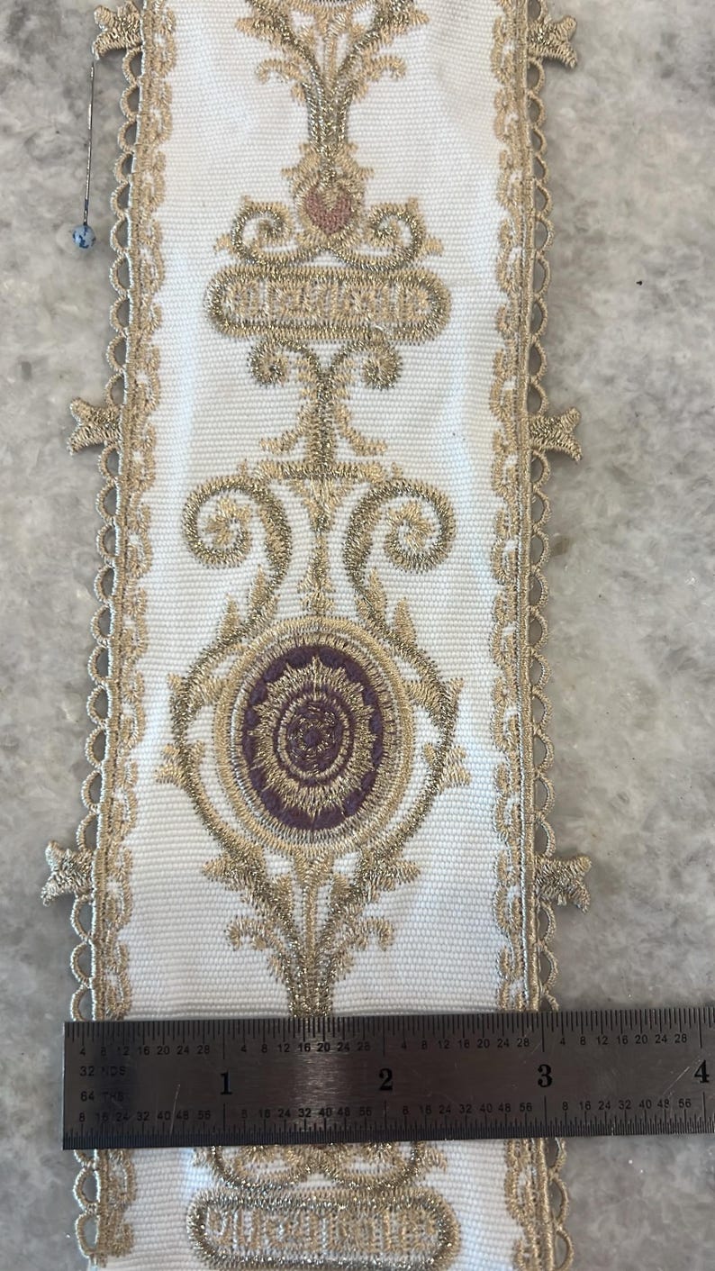 May include: A white fabric trim featuring intricate gold metallic embroidery and a central purple circular motif. The trim has scalloped edges and is positioned next to a metal ruler showing a width of approximately 3.5 inches.