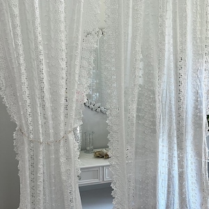 May include: White lace curtains with intricate floral and geometric patterns. The curtains are semi-sheer, allowing light to filter through. A decorative chain with small embellishments hangs between the curtains. The curtains frame a white vanity with a mirror.