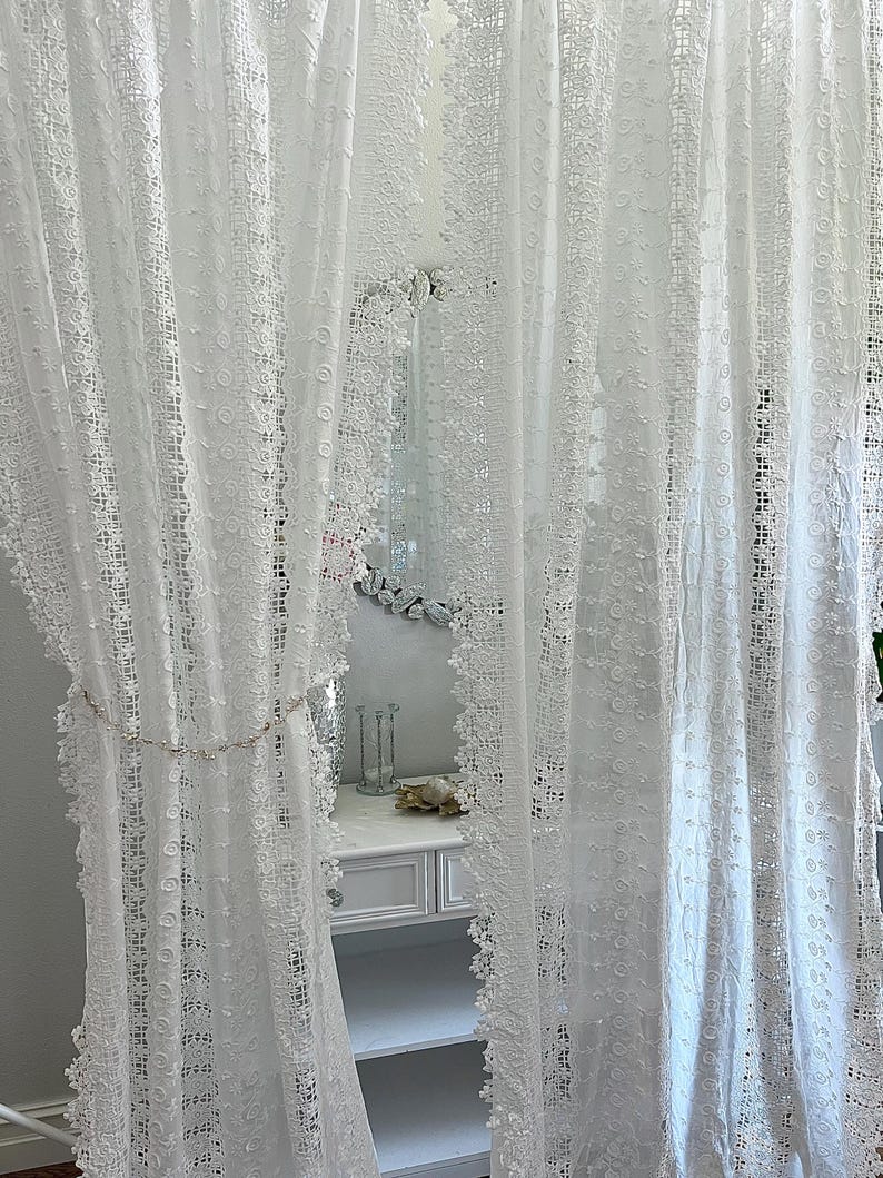 May include: White lace curtains with intricate floral and geometric patterns. The curtains are semi-sheer, allowing light to filter through. A decorative chain with small embellishments hangs between the curtains. The curtains frame a white vanity with a mirror.