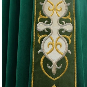 May include: Close-up of a green liturgical vestment with an embroidered border. The border features a white central design with gold and silver accents. The fabric has a soft, velvet-like texture.