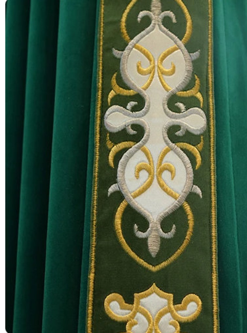 May include: Close-up of a green liturgical vestment with an embroidered border. The border features a white central design with gold and silver accents. The fabric has a soft, velvet-like texture.