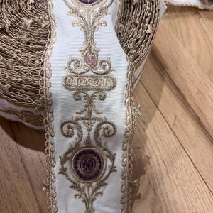 May include: A roll of white fabric trim featuring an intricate embroidered pattern in metallic gold thread and circular purple accents. The trim has scalloped edges and a repeating vertical design of ornate floral and geometric motifs.