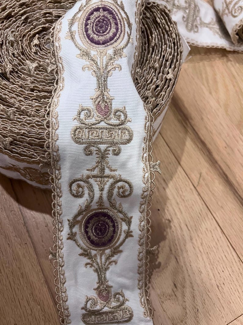 May include: A roll of white fabric trim featuring an intricate embroidered pattern in metallic gold thread and circular purple accents. The trim has scalloped edges and a repeating vertical design of ornate floral and geometric motifs.