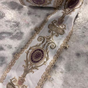 May include: A white ribbon featuring an intricate gold metallic embroidered pattern with repeating purple circular motifs. The edges of the ribbon are finished with a delicate gold scalloped lace trim, creating a decorative border along both sides.
