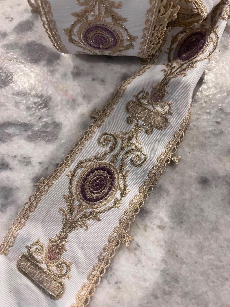 May include: A white ribbon featuring an intricate gold metallic embroidered pattern with repeating purple circular motifs. The edges of the ribbon are finished with a delicate gold scalloped lace trim, creating a decorative border along both sides.