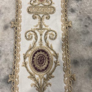 May include: A white fabric trim featuring an intricate embroidered pattern in metallic gold thread and deep burgundy accents. The design includes symmetrical scrollwork and circular motifs, finished with a decorative scalloped gold lace edge.
