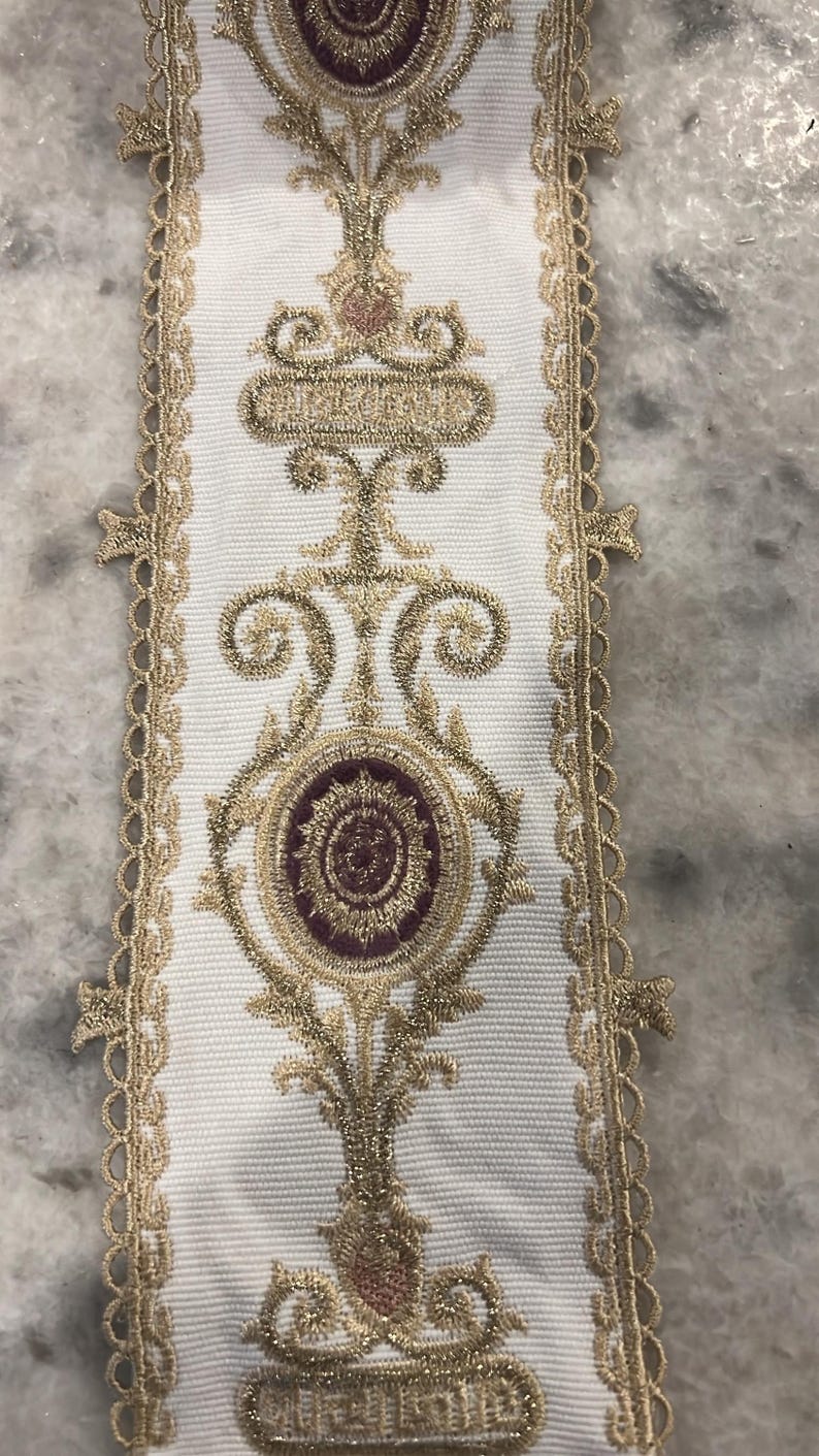 May include: A white fabric trim featuring an intricate embroidered pattern in metallic gold thread and deep burgundy accents. The design includes symmetrical scrollwork and circular motifs, finished with a decorative scalloped gold lace edge.