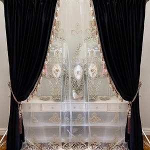 Pu&ograve; includere: Eleganti tende in velluto nero con fermatende a nappe dorate incorniciano una tenda bianca trasparente con ricami dorati. Le tende sono appese a un'asta dorata, creando un aspetto lussuoso e sofisticato.