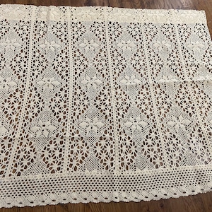 Op de afbeelding: Crèmekleurig, open geweven gehaakt textiel. Het ingewikkelde patroon heeft bloemen en geometrische ontwerpen, met een geschulpte rand. Het textiel is waarschijnlijk een tafelkleed of decoratief stuk, dat gedetailleerd vakmanschap laat zien.