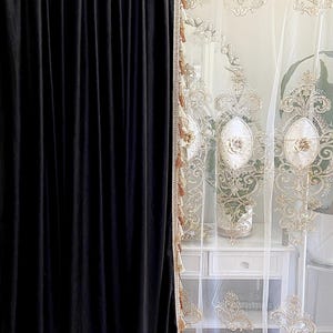 Pu&ograve; includere: Pannello di tenda in velluto nero accanto a una tenda bianca trasparente con ricami floreali dorati. La tenda trasparente ha un bordo con nappe decorative. Un comodino bianco &egrave; visibile dietro la tenda trasparente.