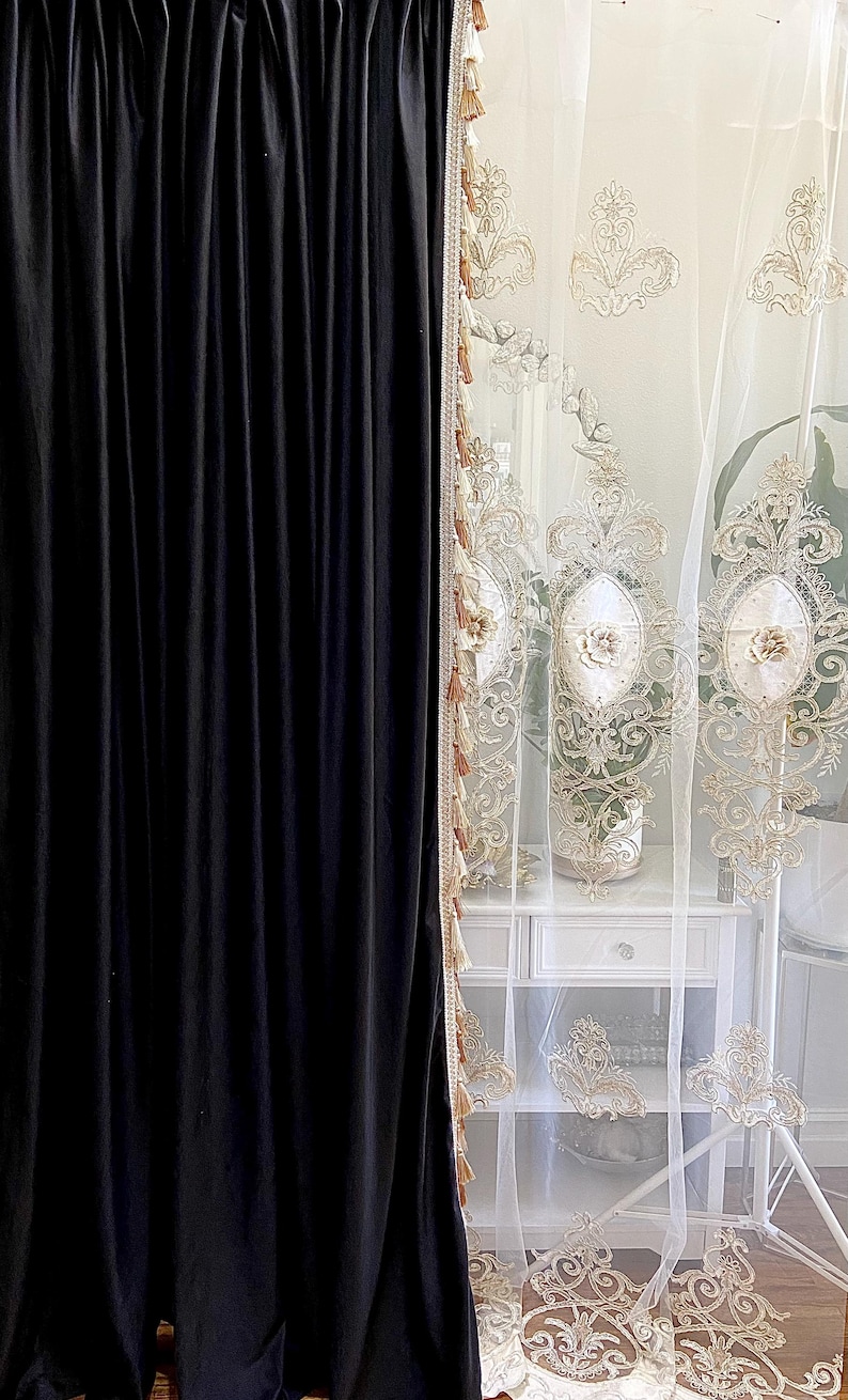Pu&ograve; includere: Pannello di tenda in velluto nero accanto a una tenda bianca trasparente con ricami floreali dorati. La tenda trasparente ha un bordo con nappe decorative. Un comodino bianco &egrave; visibile dietro la tenda trasparente.