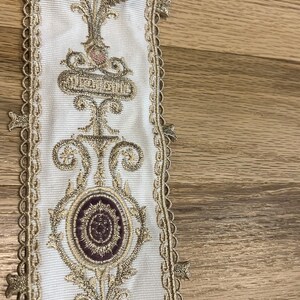 May include: A white fabric trim featuring an intricate embroidered pattern in metallic gold thread and deep purple accents. The design includes repeating circular motifs and scrollwork, finished with a scalloped gold edge along both sides.