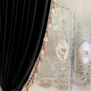 Pu&ograve; includere: Tenda in velluto nero con finiture di perline e nappe decorative. Tenda bianca trasparente con ricamo floreale. Le tende sono appese davanti a un com&ograve; bianco.