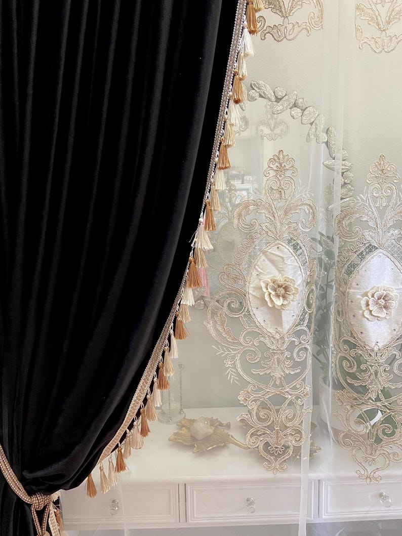 Pu&ograve; includere: Tenda in velluto nero con finiture di perline e nappe decorative. Tenda bianca trasparente con ricamo floreale. Le tende sono appese davanti a un com&ograve; bianco.