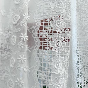 May include: Close-up of white lace curtains with floral and geometric patterns. The curtains feature embroidered flowers, leaves, and delicate openwork designs. The intricate details create a textured and elegant look, perfect for home decor.