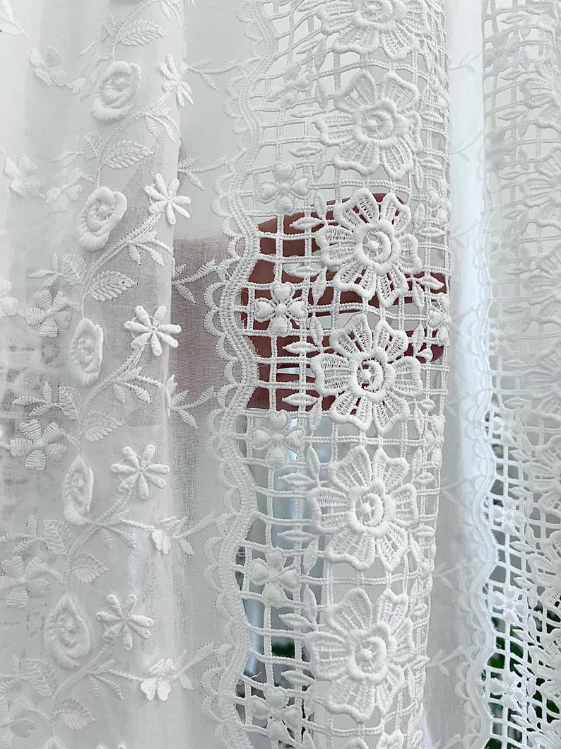 May include: Close-up of white lace curtains with floral and geometric patterns. The curtains feature embroidered flowers, leaves, and delicate openwork designs. The intricate details create a textured and elegant look, perfect for home decor.
