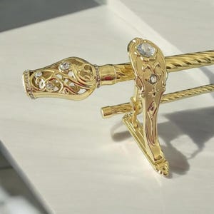 May include: Ornate gold-colored curtain rod with decorative finials and a matching clip. The finials feature intricate openwork designs and are embellished with clear, sparkling crystals. The rod itself has a twisted rope-like texture.