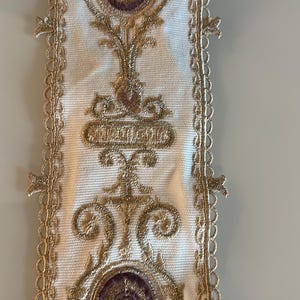 May include: A white fabric trim featuring intricate gold metallic embroidery and circular purple accents. The vertical strip has a scalloped lace edge on both sides, with repeating symmetrical patterns of gold thread and central purple circular motifs.