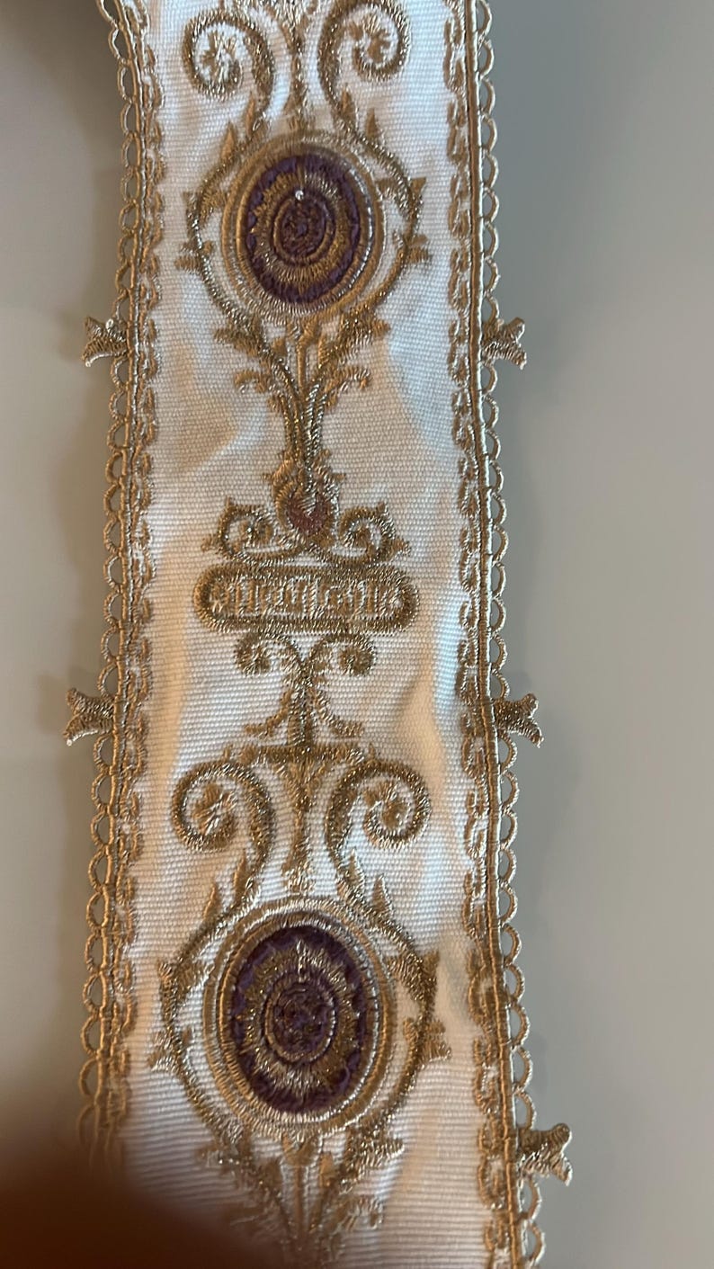 May include: A white fabric trim featuring intricate gold metallic embroidery and circular purple accents. The vertical strip has a scalloped lace edge on both sides, with repeating symmetrical patterns of gold thread and central purple circular motifs.
