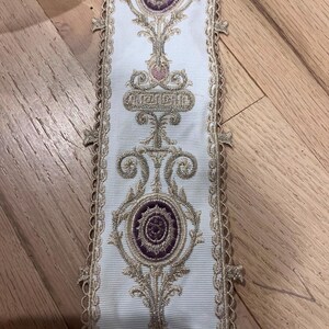 May include: A white fabric trim featuring an intricate embroidered pattern in metallic gold thread and deep purple accents. The design includes repeating circular motifs and scrollwork, finished with a scalloped, decorative gold-toned edge.