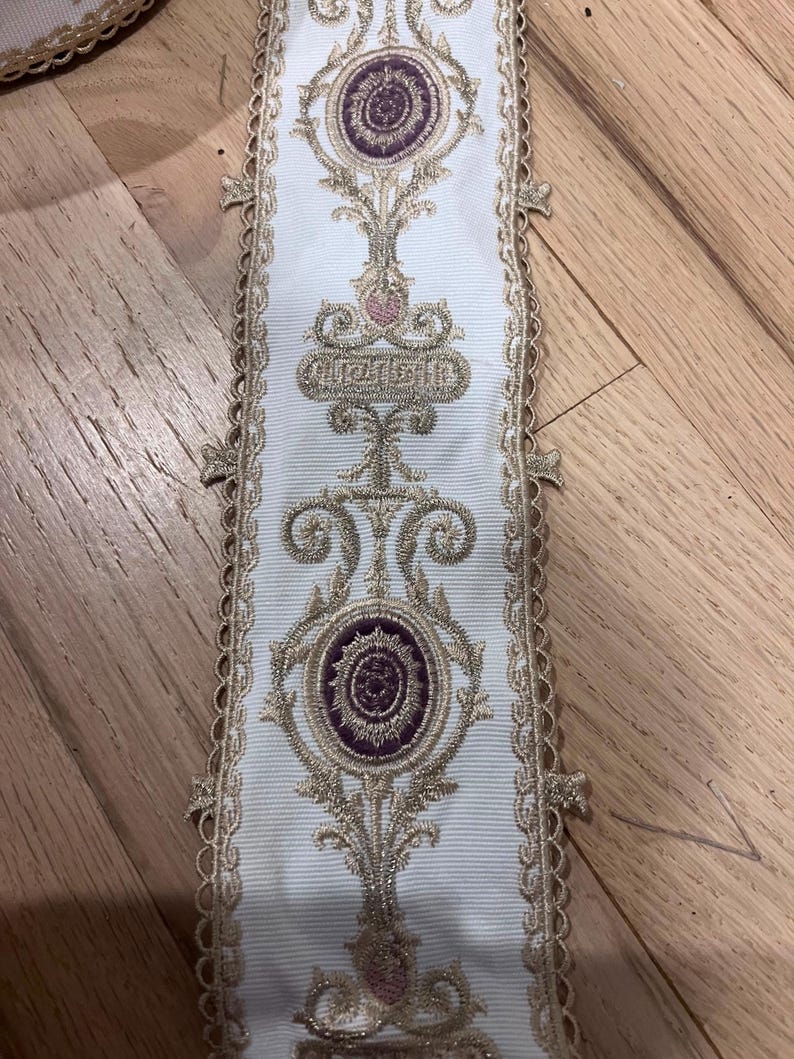 May include: A white fabric trim featuring an intricate embroidered pattern in metallic gold thread and deep purple accents. The design includes repeating circular motifs and scrollwork, finished with a scalloped, decorative gold-toned edge.