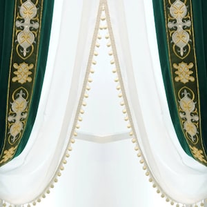 May include: Dark green velvet curtains with gold and cream embroidered borders. Sheer white curtains with cream pom-pom trim are layered behind the velvet drapes, creating a luxurious window dressing.