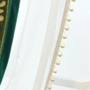 May include: White sheer fabric with a cream trim featuring small, round pom-poms. A section of green fabric with gold embroidery is visible on the left. The design suggests a curtain or decorative textile.