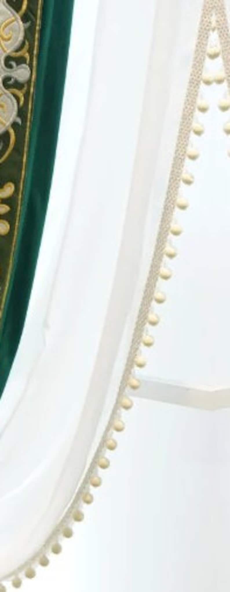 May include: White sheer fabric with a cream trim featuring small, round pom-poms. A section of green fabric with gold embroidery is visible on the left. The design suggests a curtain or decorative textile.