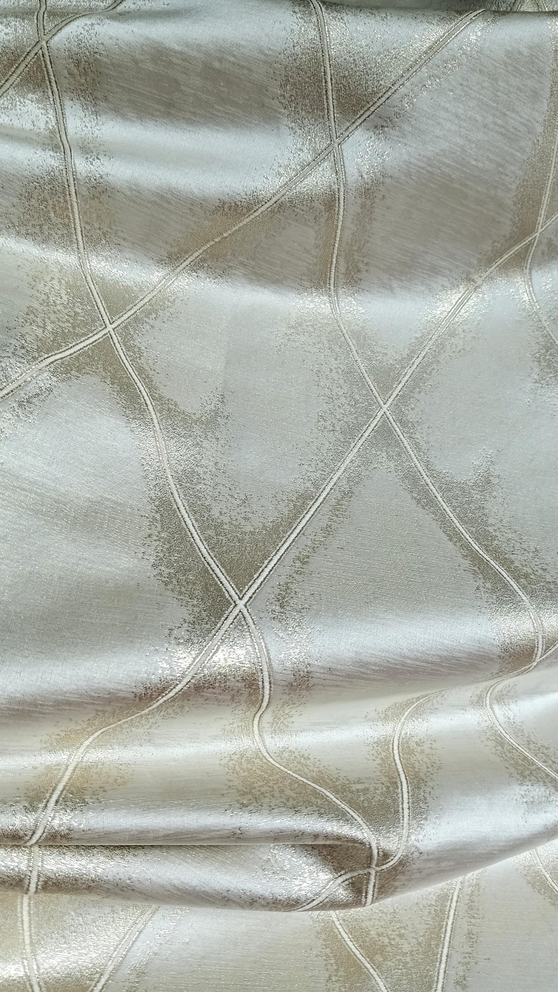 May include: Close-up of a shimmering, textured fabric in shades of silver and beige. The material features a repeating pattern of intersecting lines, creating a diamond-like grid design. The fabric has a subtle sheen.