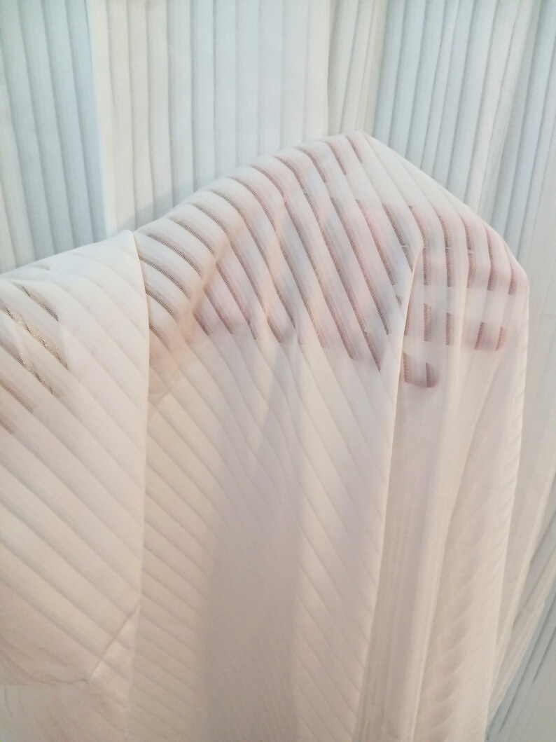 May include: A sheer white fabric with diagonal stripes is held up, revealing a hand through the material. The fabric has a pleated texture, and the background is a similar white, pleated material.
