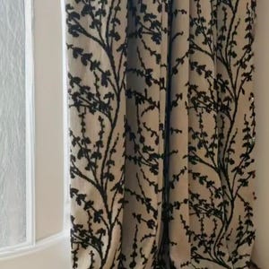 May include: Beige and black patterned curtains. The curtains have a repeating design of black branches with leaves against a beige backdrop. They hang in front of a white window frame and a light-coloured floor.