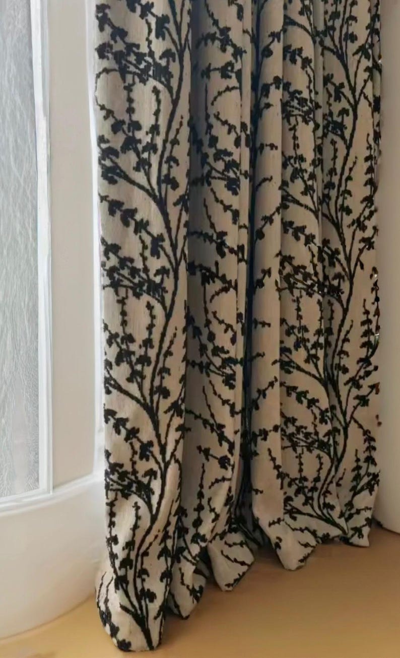 May include: Beige and black patterned curtains. The curtains have a repeating design of black branches with leaves against a beige backdrop. They hang in front of a white window frame and a light-coloured floor.