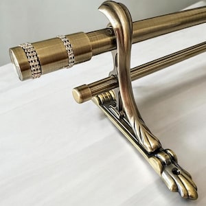 May include: Antique brass-colored curtain rod with decorative elements. The rod features a cylindrical end with two rows of sparkling rhinestones. The support bracket has a curved, ornate design. The rod is designed to hold curtains.