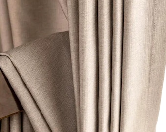 May include: Close-up of beige fabric curtains with vertical folds. The fabric has a subtle texture and a neutral color, creating a soft and elegant look. The curtains are partially draped, revealing a glimpse of a wooden frame.