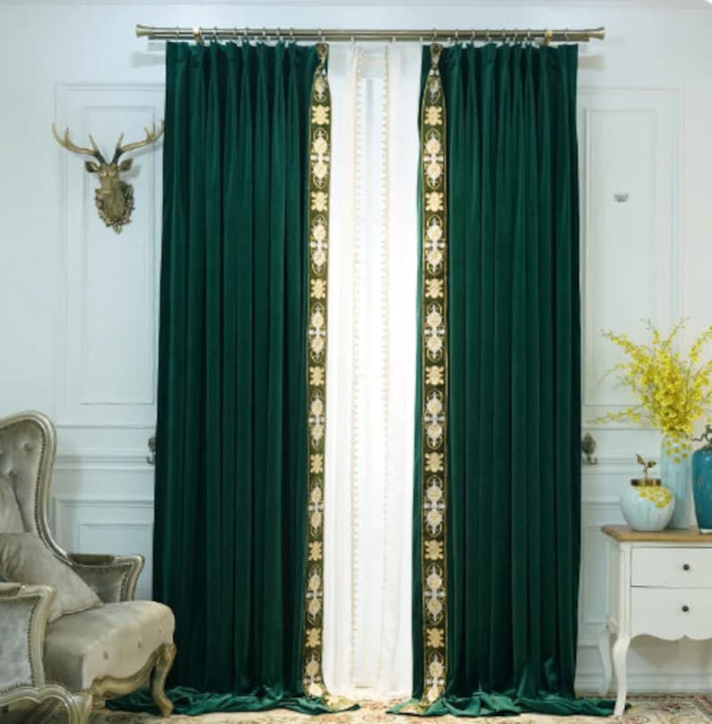 May include: Dark green velvet curtains with gold embroidered detailing. The curtains are suspended from a gold rail and frame a white sheer curtain. The room has a classic, elegant style.